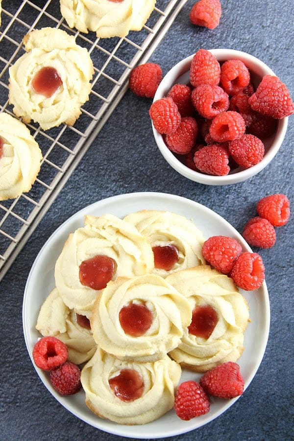 Raspberry Butter Cookies are sweet, simple, and perfect for gifting. Light, crisp butter cookies with a dollop of raspberry jam are great with a cup of tea! Raspberry Butter Cookies on blue background