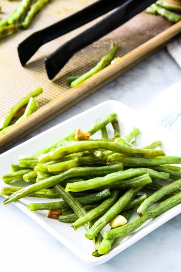These easy sheet pan Roasted Green Beans are the simplest side dish ever! With just 4 ingredients and 20 minutes they will be cooked to perfection! Sheet Pan Roasted Green Beans on white plate with sheet pan in background