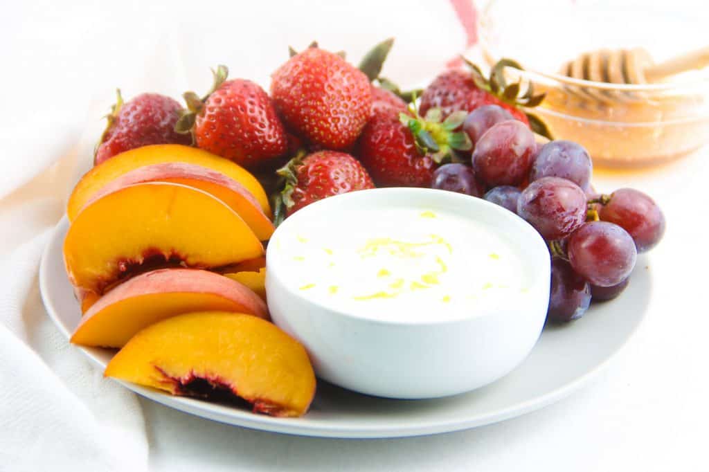 This Easy Honey Yogurt Dip is flavored with honey, lemon zest and vanilla! Top this homemade with granola or serve with fresh fruits! Honey Yogurt Dip in white bowl on grey plate with fresh fruit.