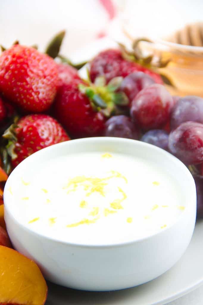 This Easy Honey Yogurt Dip is flavored with honey, lemon zest and vanilla! Top this homemade with granola or serve with fresh fruits! Honey Yogurt Dip in white bowl with fruit in background.