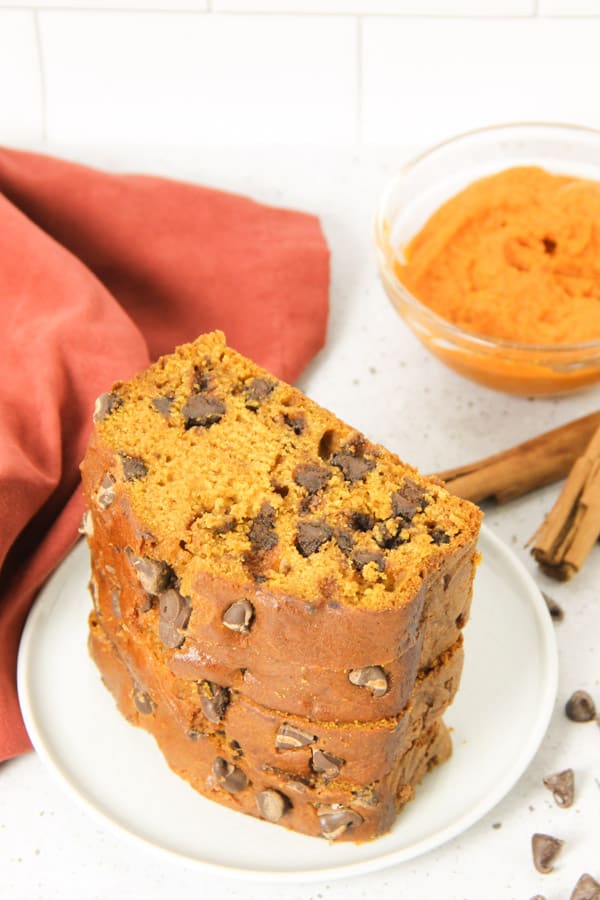 Delicious and easy Chocolate Chip Pumpkin Bread! Moist and tender, this bread is filled with warm spices and rich chocolate! Chocolate Chip Pumpkin Bread on white plate with pumpkin in a glass bowl