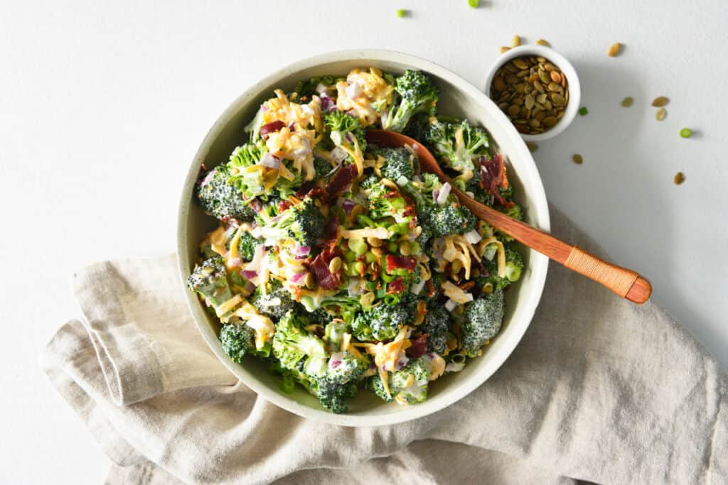 This Easy Broccoli Salad is made with bacon, cheddar cheese and a creamy mayonnaise sauce! Easy Broccoli Salad in white bowl with spoon.