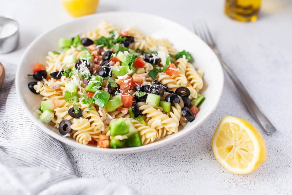 Easy Greek Pasta Salad served cold with a homemade vinaigrette. With fresh vegetables topped with feta cheese! Greek Pasta Salad in white bowl.