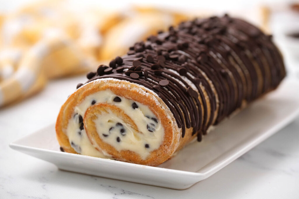 This Chocolate Chip Pumpkin Roll is a fun twist on a classic pumpkin roll! With a fluffy cream cheese filling and mini chocolate chips! Chocolate Chip Pumpkin Roll on white plate drizzled with chocolate.