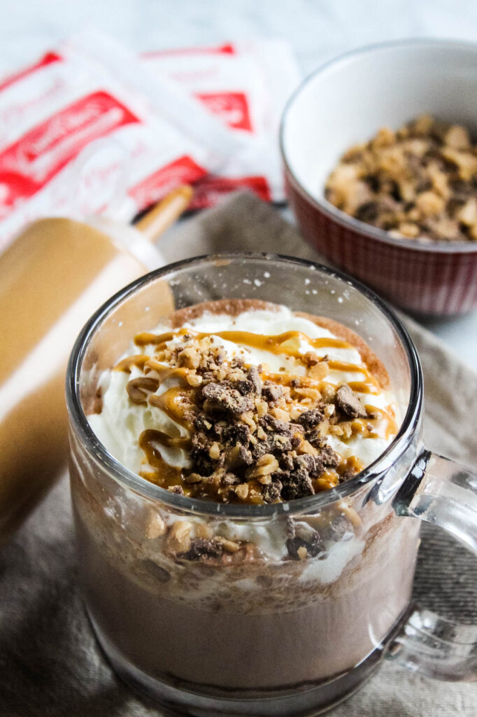 Warm up this season with this easy to make Peanut Butter Hot Chocolate topped with toffee and whipped cream. Perfect for an afternoon treat! Peanut Butter Hot Chocolate in glass mug with toppings in background.