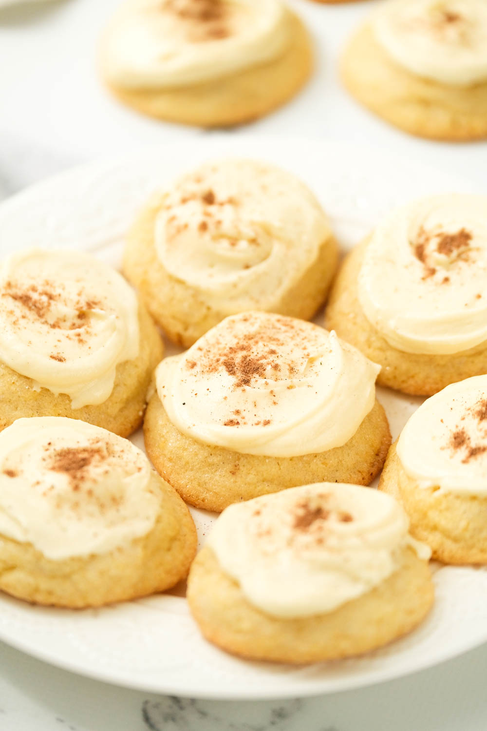 These frosted eggnog cookies are the perfect treat for eggnog lovers! With a touch of nutmeg and topped with eggnog buttercream frosting! soft eggnog cookies on white plate.