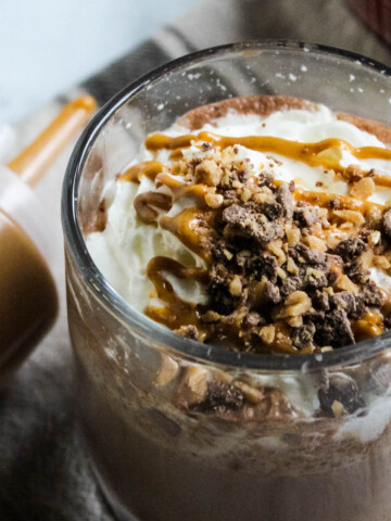 Peanut Butter Hot Chocolate in glass mug topped with whipped cream, peanut butter, and toffee bits.